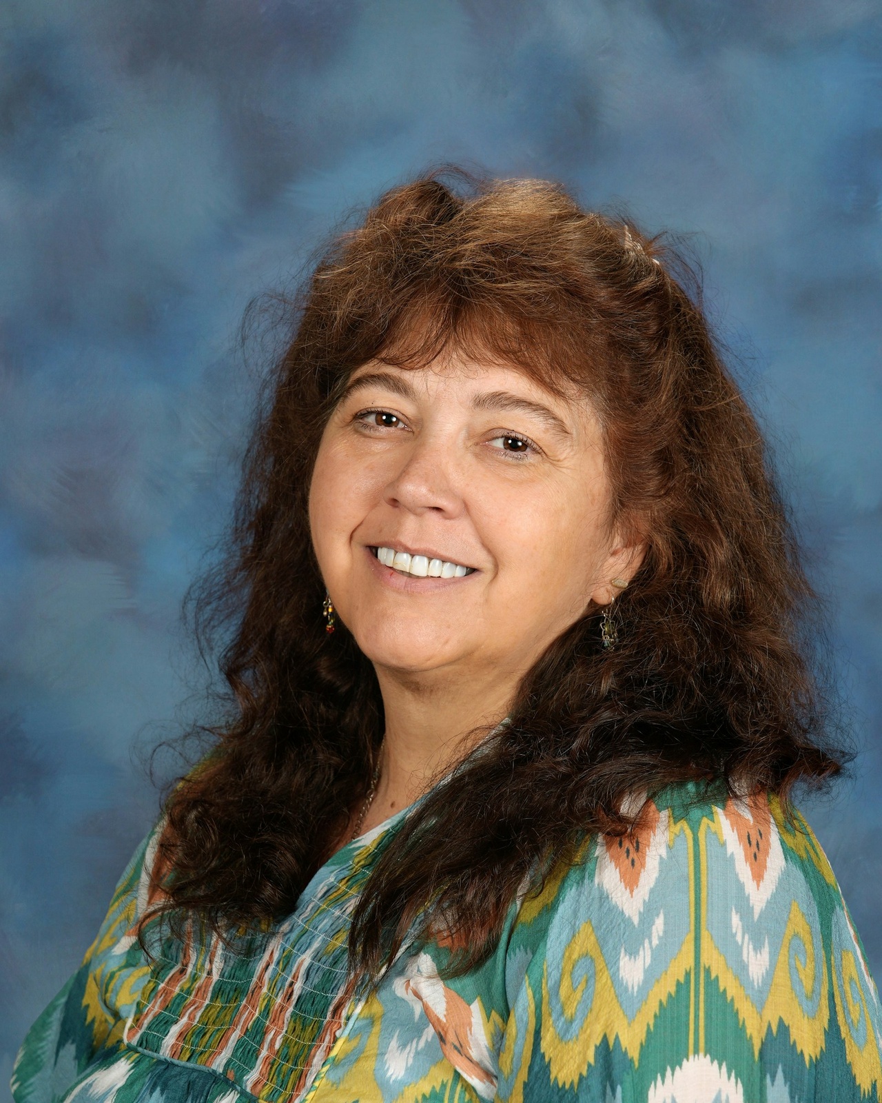 A woman with long brown hair smiles warmly against a soft blue backdrop. She's wearing a colorful patterned top with an inviting expression.